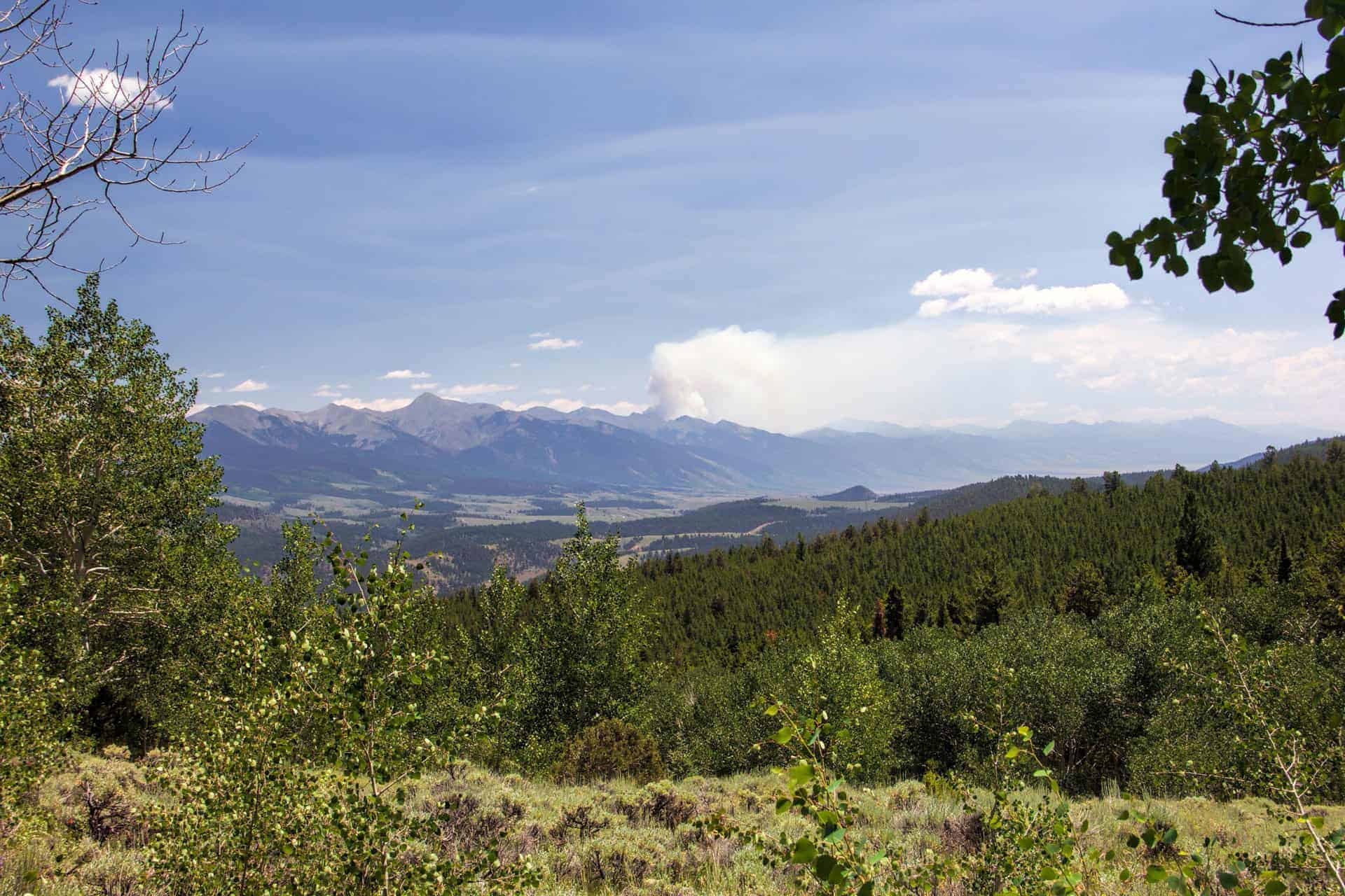 A hazy mountain valley view with a large plume of wildfire smoke rising on the horizon