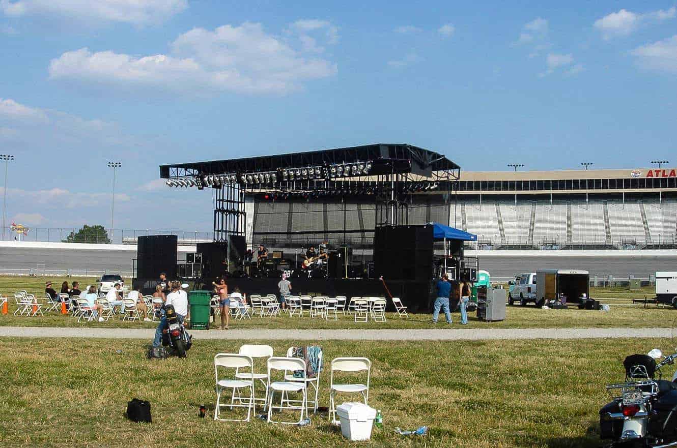 A band performing on an outdoor stage at the Atlanta Motor Speedway's motorcycle rally, with empty grandstands and folding chairs on the infield grass