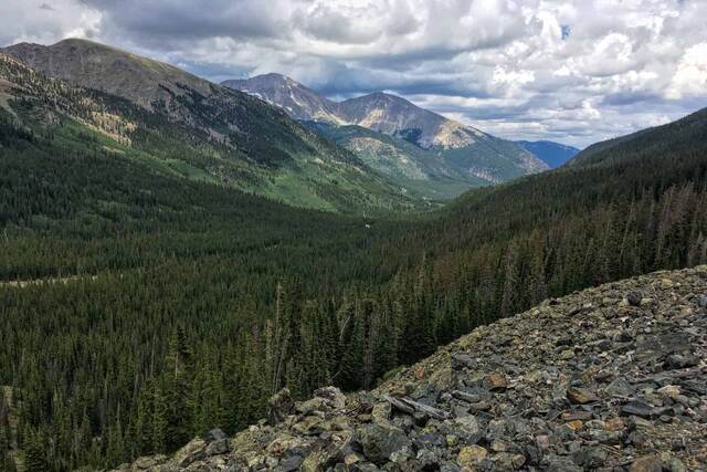A view down a forested valley toward layered mountain peaks under a partly cloudy sky