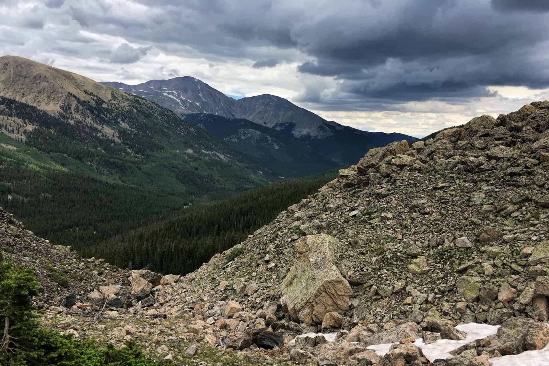 A view down a forested valley toward distant peaks with dark storm clouds building overhead