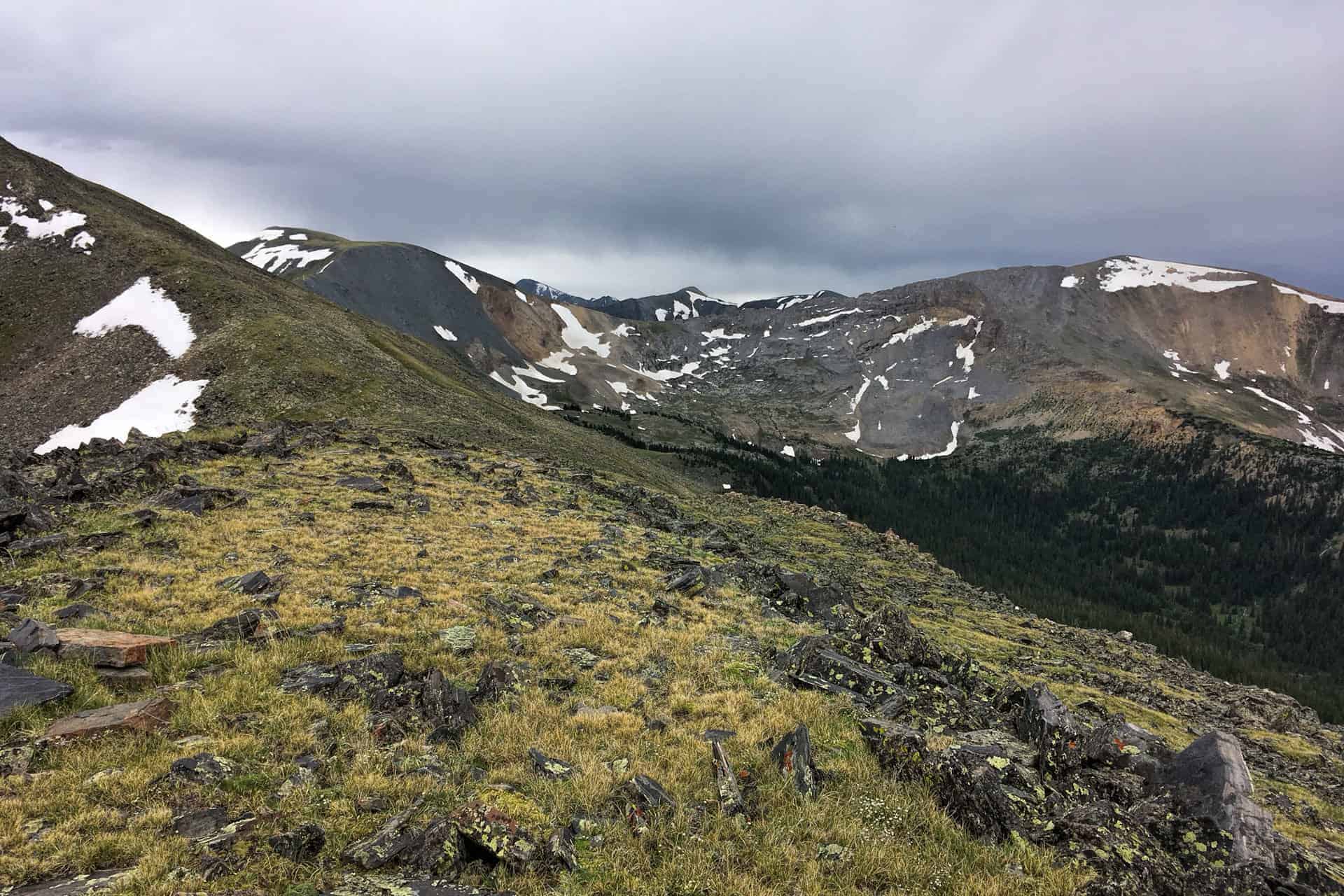 A grassy, rock-strewn alpine ridge overlooking a broad snow-streaked mountain bowl under a heavily clouded sky