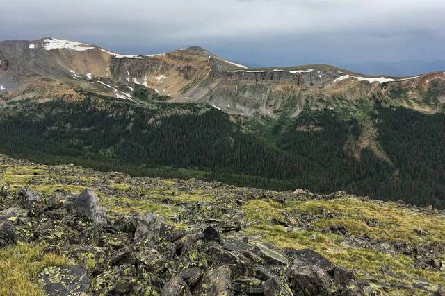 A pile of dark, lichen-covered rocks atop a grassy ridge, with a rugged snow-patched peak and stormy sky in the distance