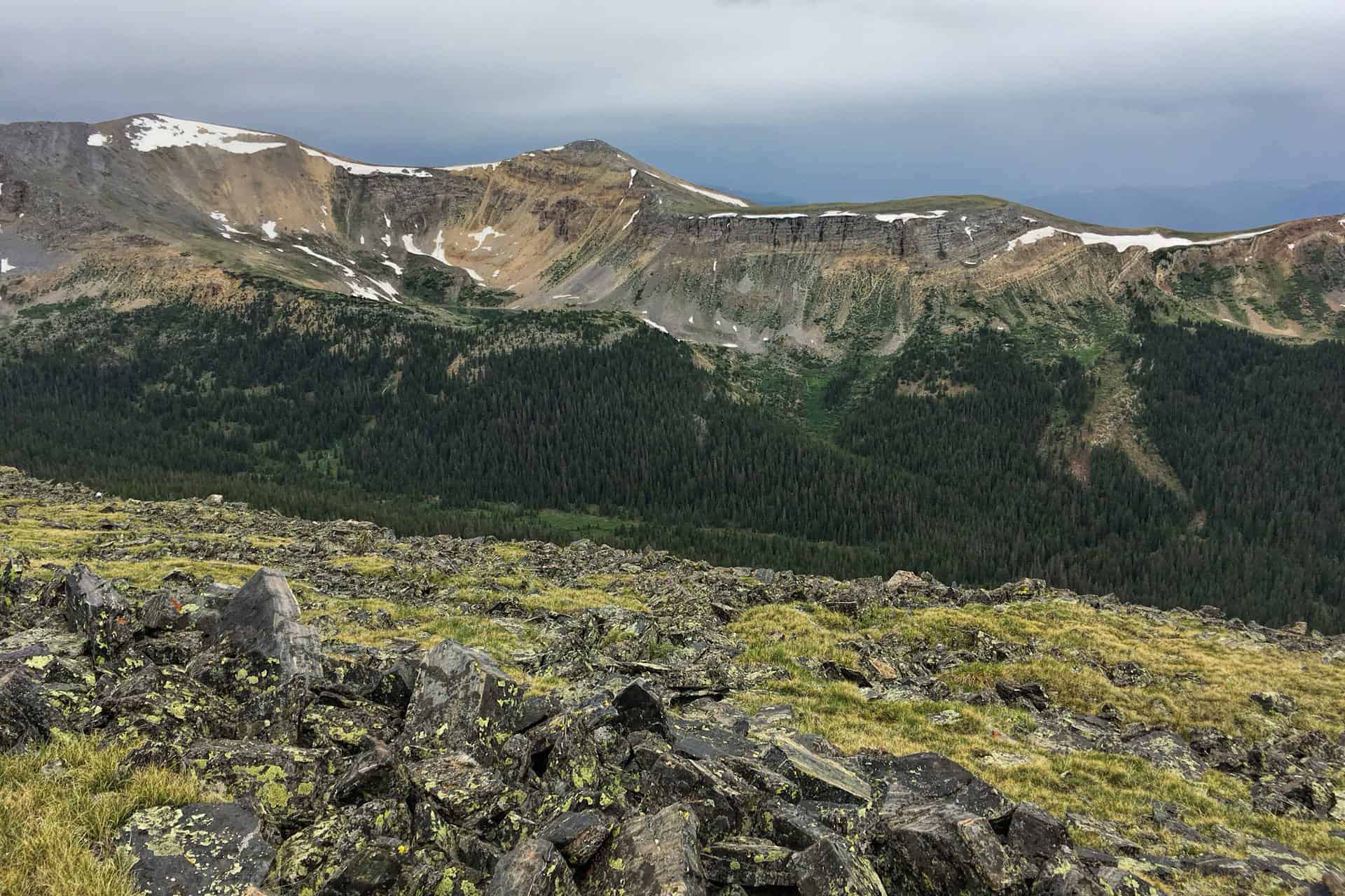 A pile of dark, lichen-covered rocks atop a grassy ridge, with a rugged snow-patched peak and stormy sky in the distance