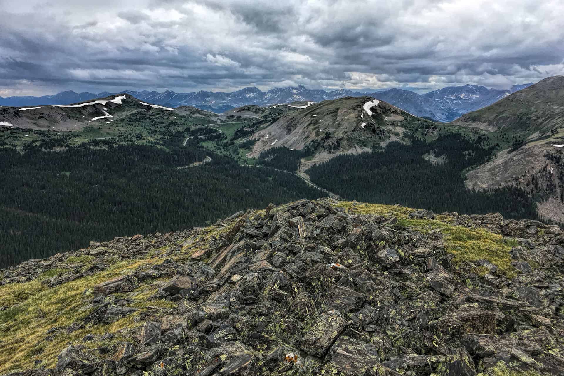 A rocky summit cairn on a grassy ridge overlooking a forested valley and distant snow-streaked mountains under a dramatic stormy sky