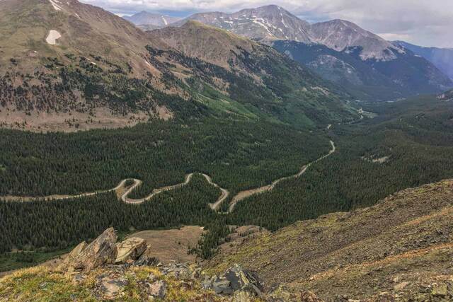 A high view down into a forested valley with a winding paved switchback road, rugged snow-patched peaks in the background