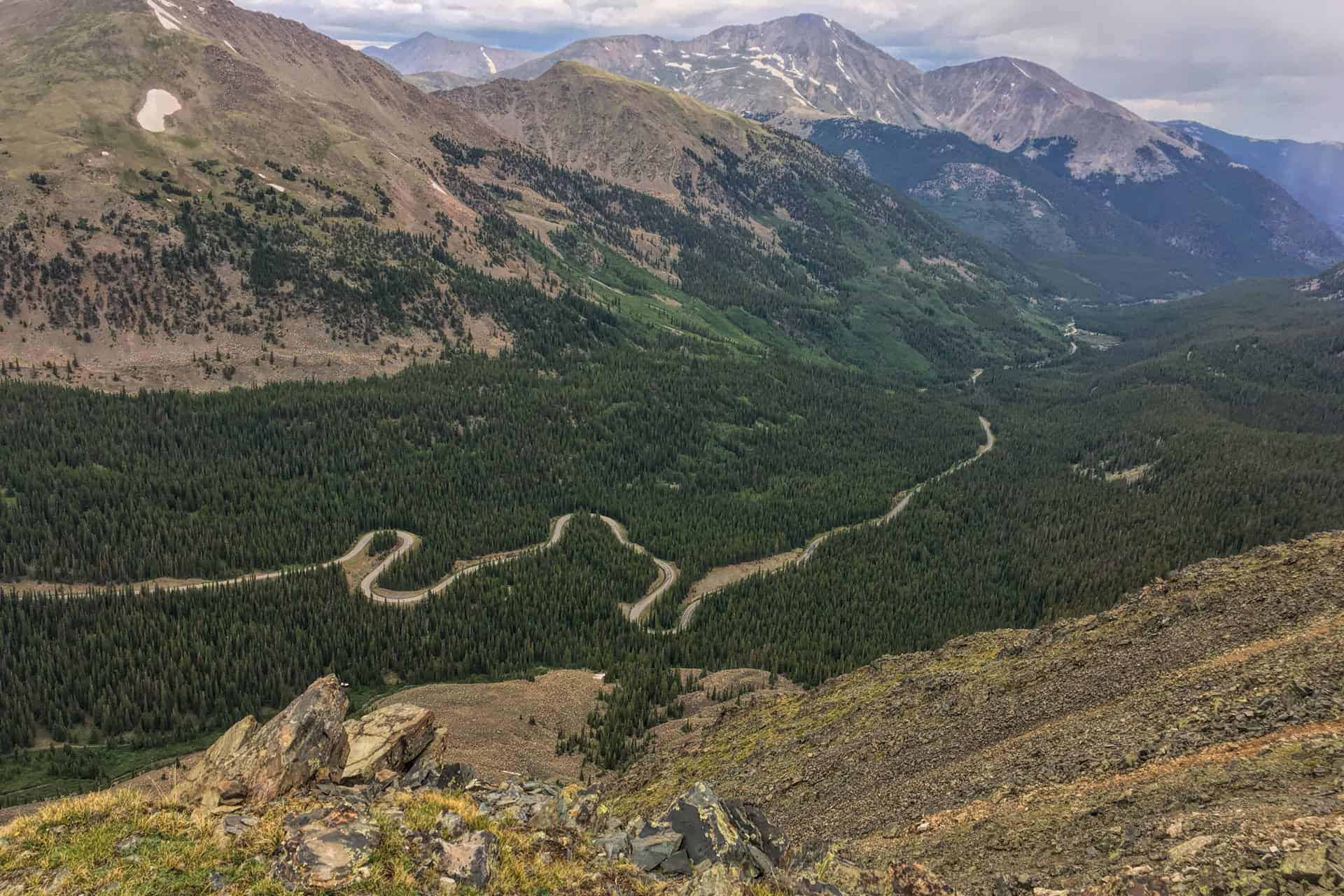 A high view down into a forested valley with a winding paved switchback road, rugged snow-patched peaks in the background