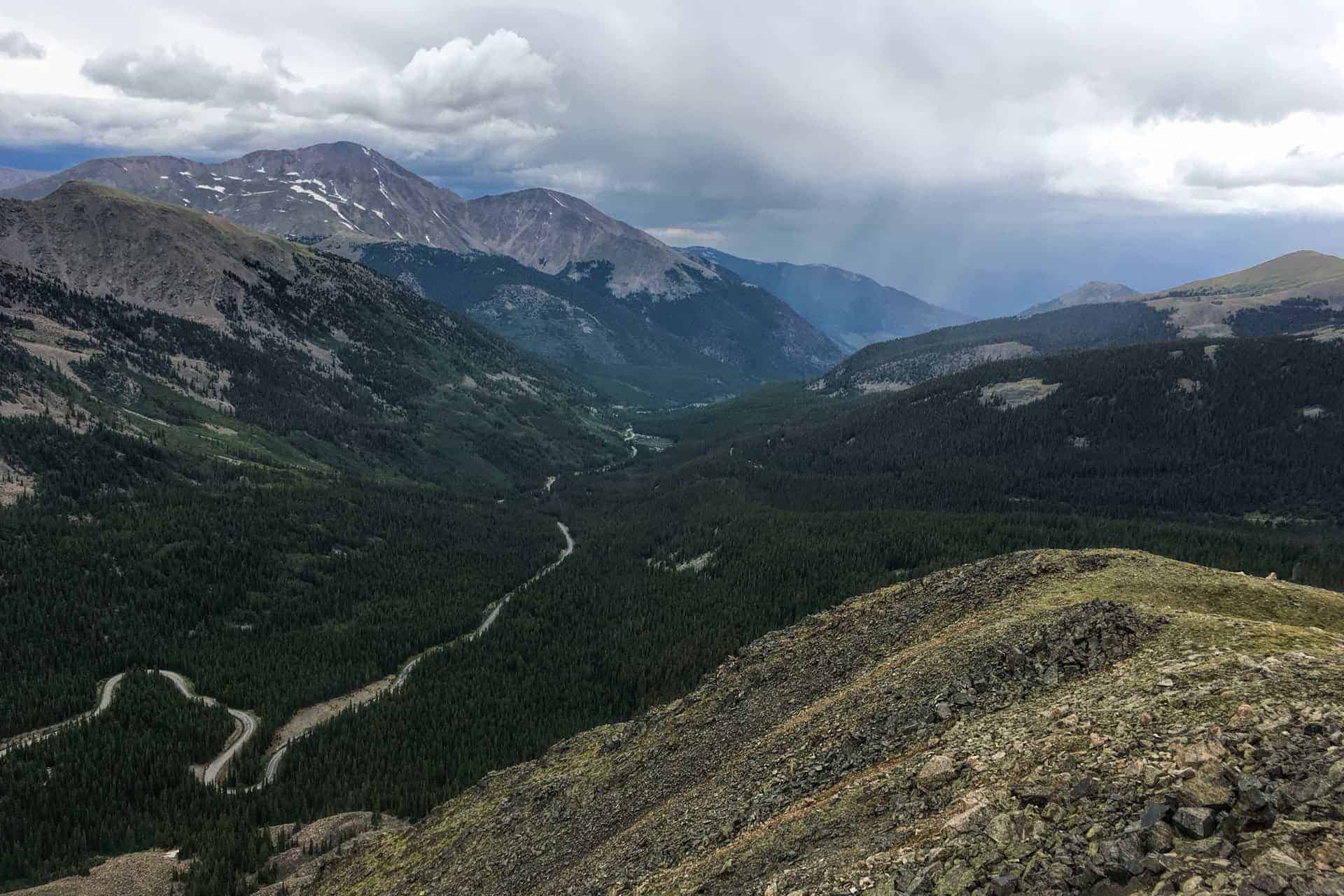 A forested valley with a winding road following a stream, mountains rising on either side and a rain shower visible in the distance