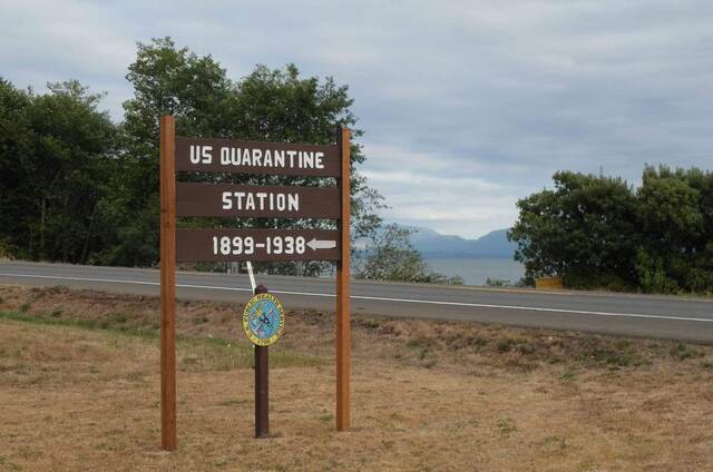A weathered wooden sign reading "US Quarantine Station 1899-1938" beside the road, with the Columbia River in the background