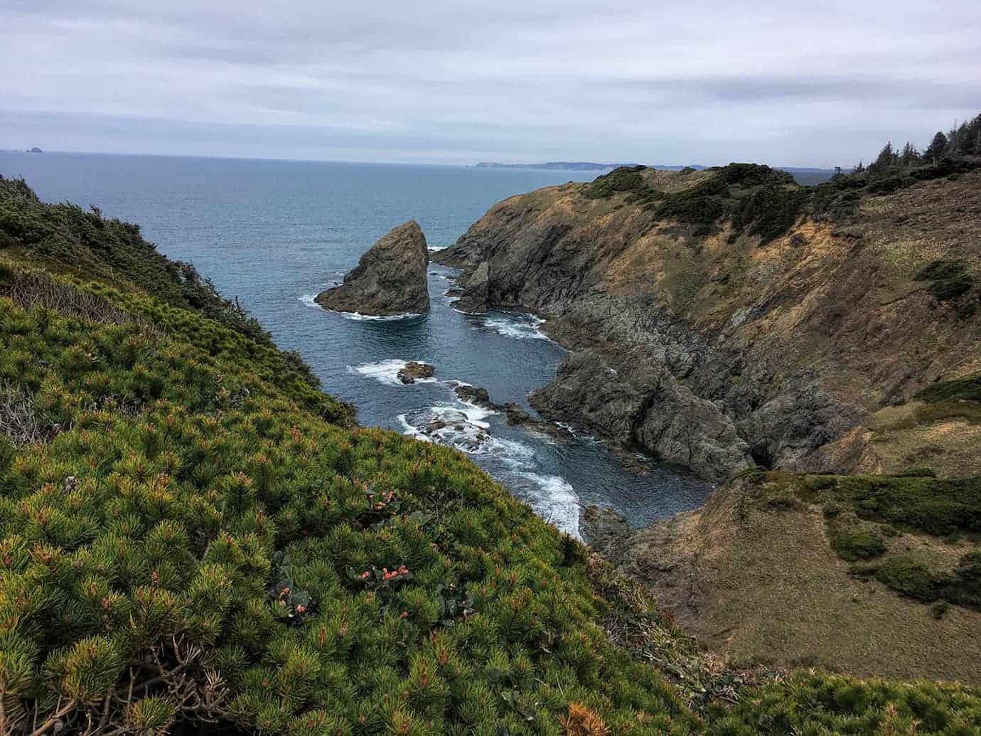 A close-up view of a coastal cove with a lone sea stack, rugged cliffs, and the ocean stretching to the horizon