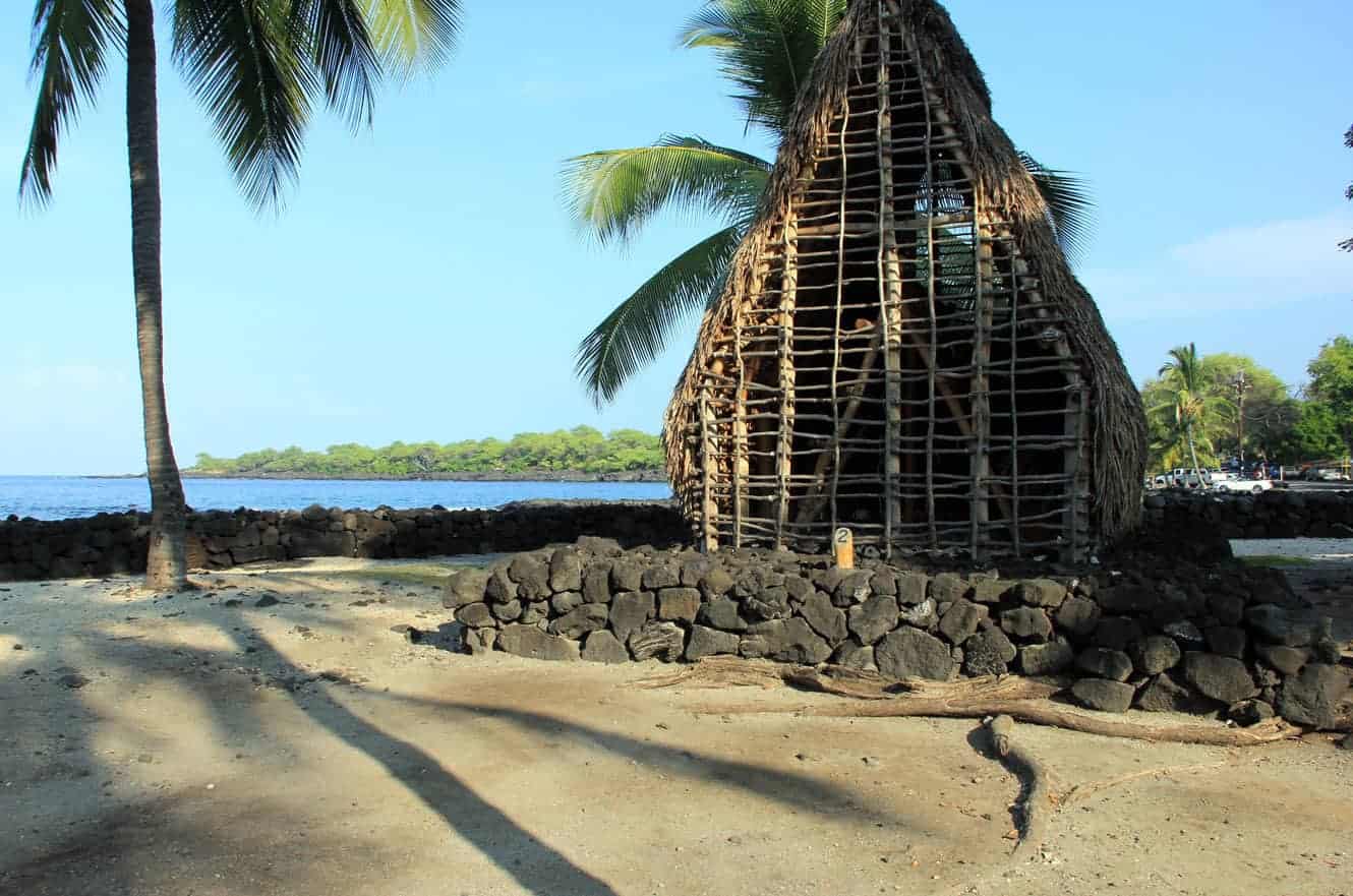 A reconstructed thatched hale on the shoreline at Pu'uhonua O Hōnaunau, framed by palm trees