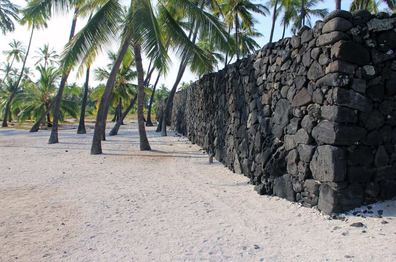 A long lava rock wall lined with palm trees along the white sand at Pu'uhonua O Hōnaunau