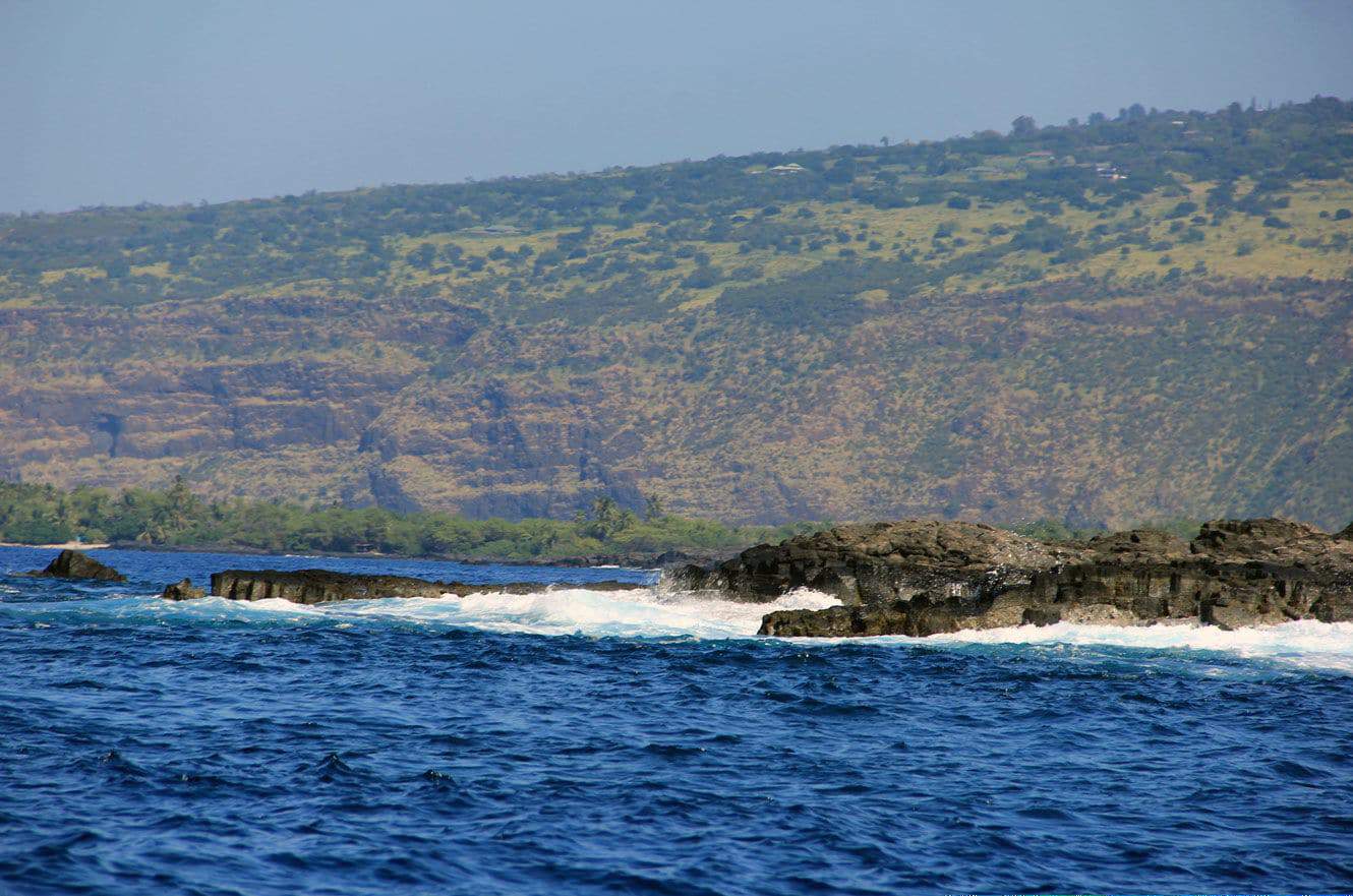A distant view of the Kona coastline's cliffs and green hillside from the water