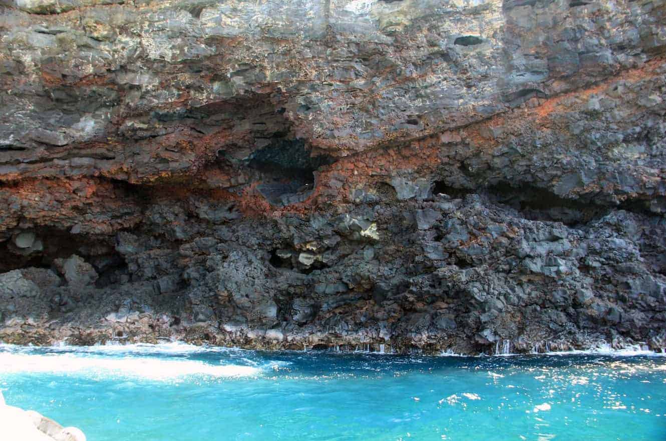 A close-up view of layered red and black lava rock forming an overhang above the water