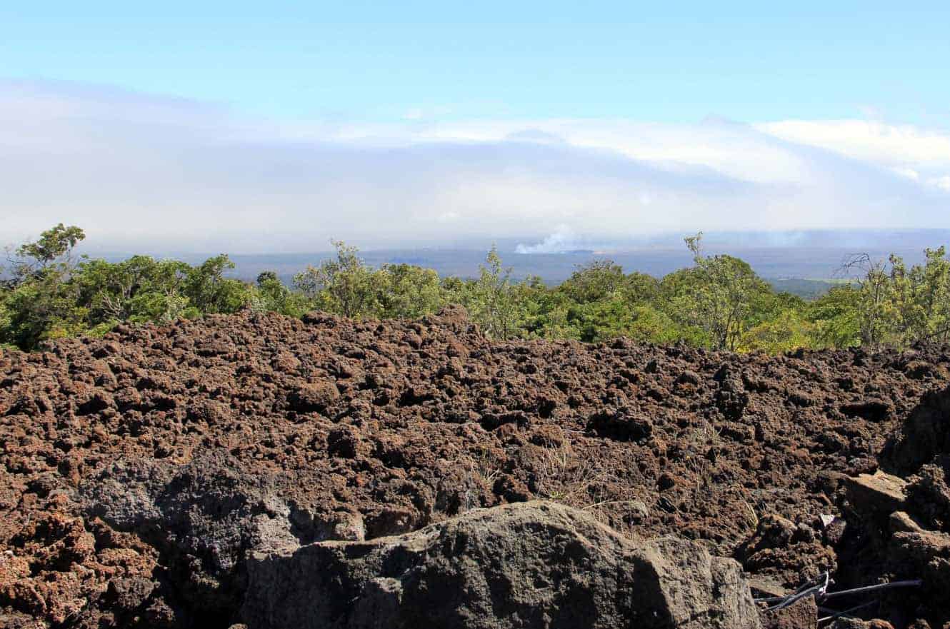 A hazy view over the lava field toward green trees, with smoke rising in the distance