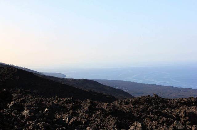 A hazy dusk view over dark lava rock toward the ocean and a distant rock outcropping
