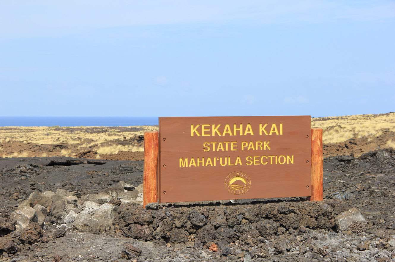 The Kekaha Kai State Park, Mahai'ula Section entrance sign amid a black lava field with the ocean beyond