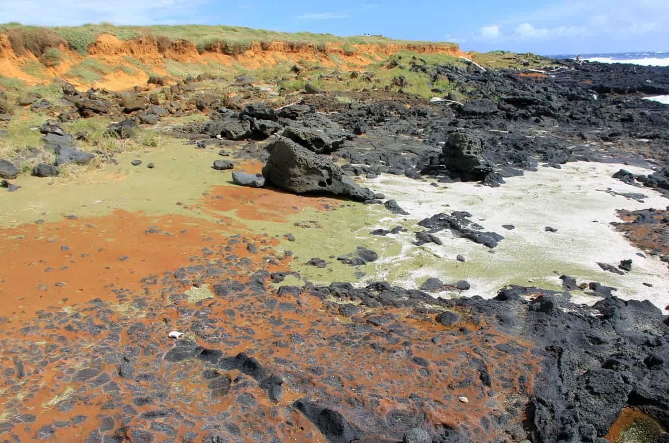 Red cinder cliffs above a rocky beach with patches of green olivine sand