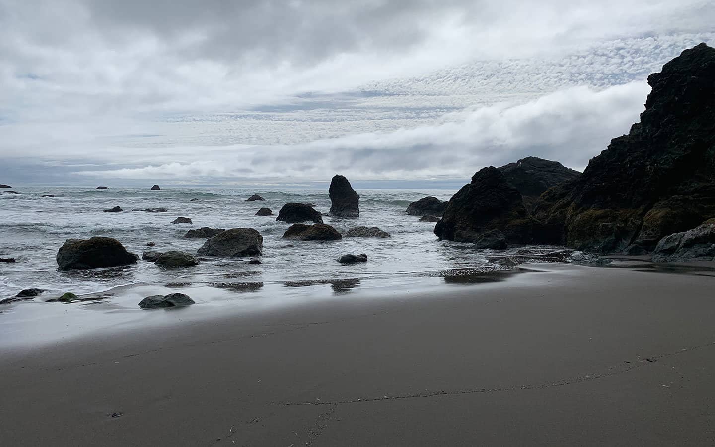 A misty, overcast beach scene with dark rock formations and a tall sea stack in the surf