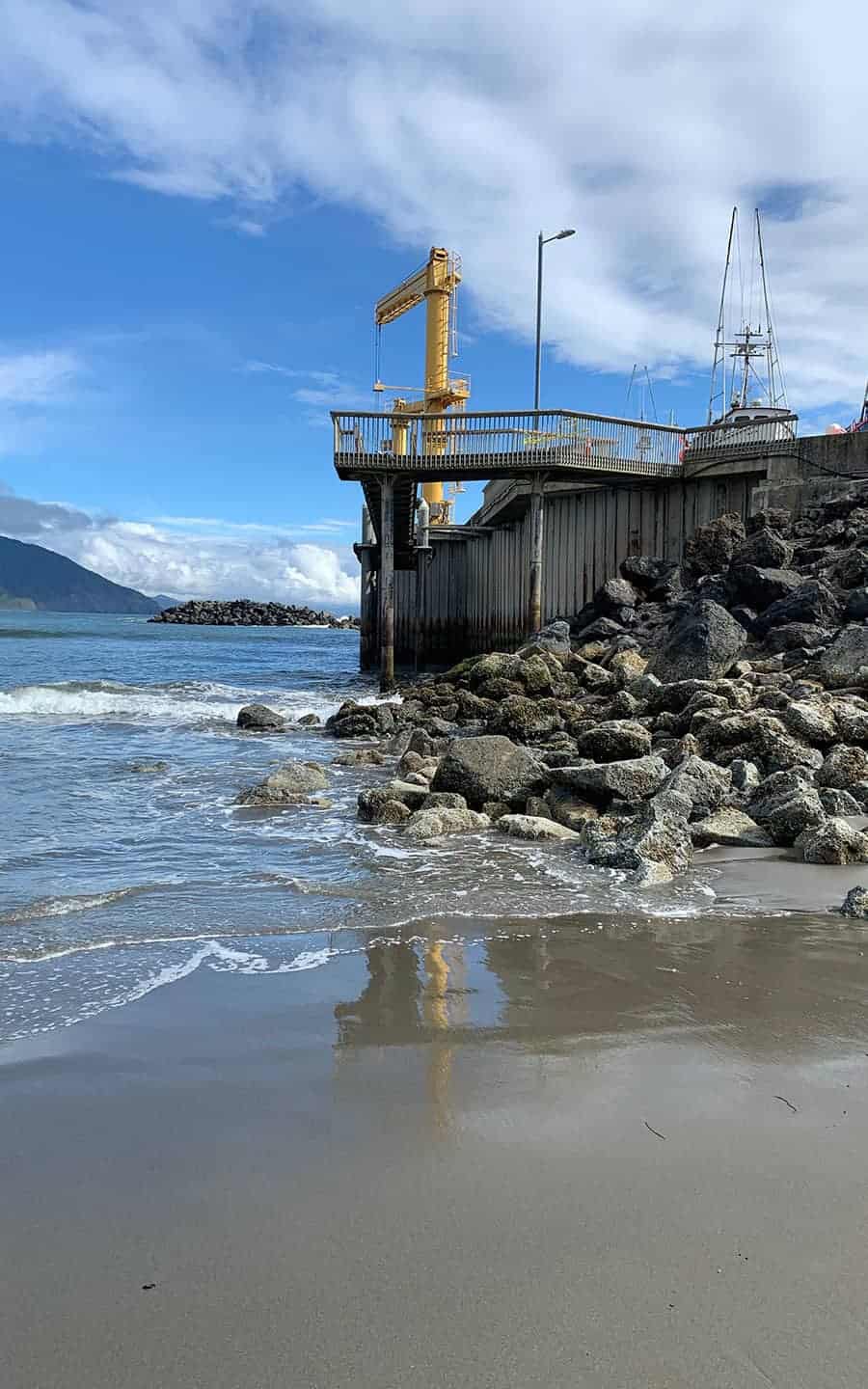 A side view of the dolly dock's wooden platform and yellow crane, used to launch fishing boats, with rocks piled along the shore