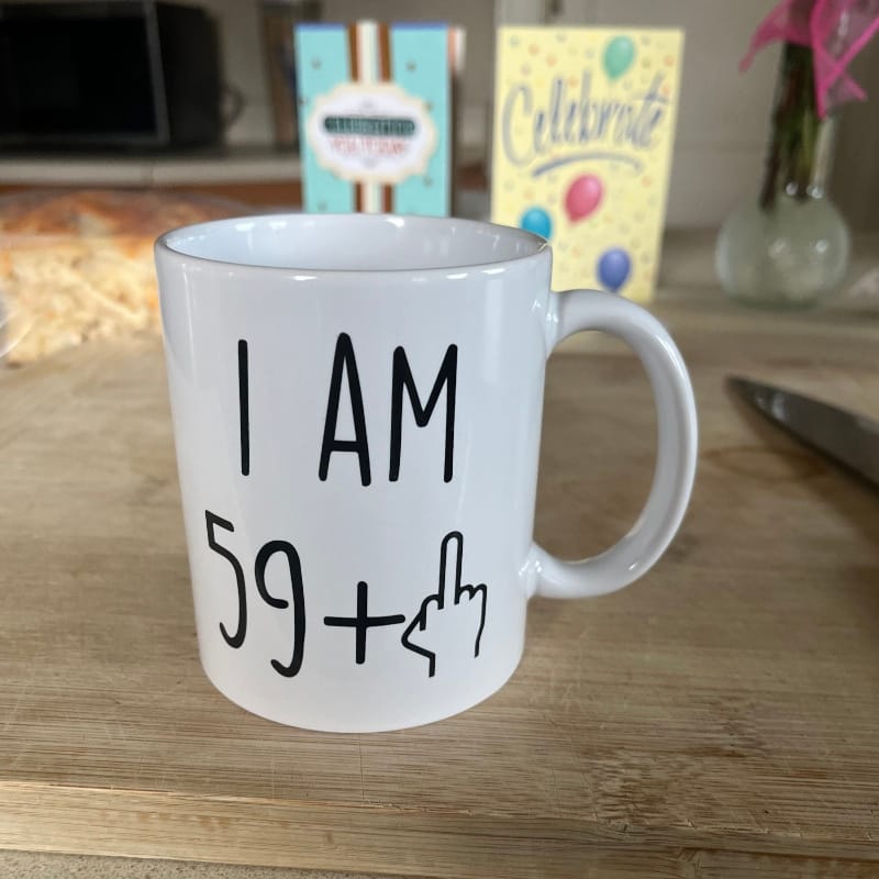 A mug reading "I AM 59+" with a hand-drawn raised middle finger, sitting on a wood counter with birthday cards and flowers in the background